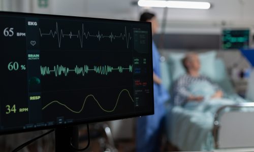 Monitor in hospital ward showing bmp from patient, laying in bed with oxymeter attached on finger, discussing with doctor and nurse about recovery treatment.